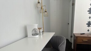 A small, modern room with a white desk, a gray chair, a coffee maker, and a bedside table with a remote control. - Grand Central Motel (Ely)