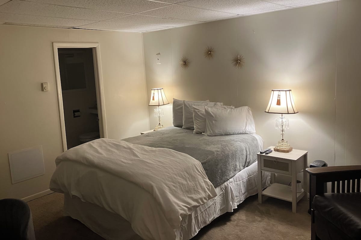 A hotel room with a bed, two bedside tables with lamps, a chair, and a door leading to a bathroom.