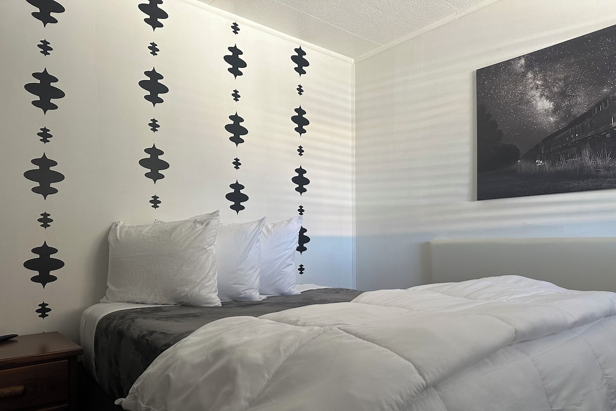 A bedroom with a bed, white bedding, a nightstand, and a wall with decorative black patterns.