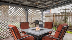 En overdækket terrasse med bålstedbord, fire stole, grill og espalierhegn.