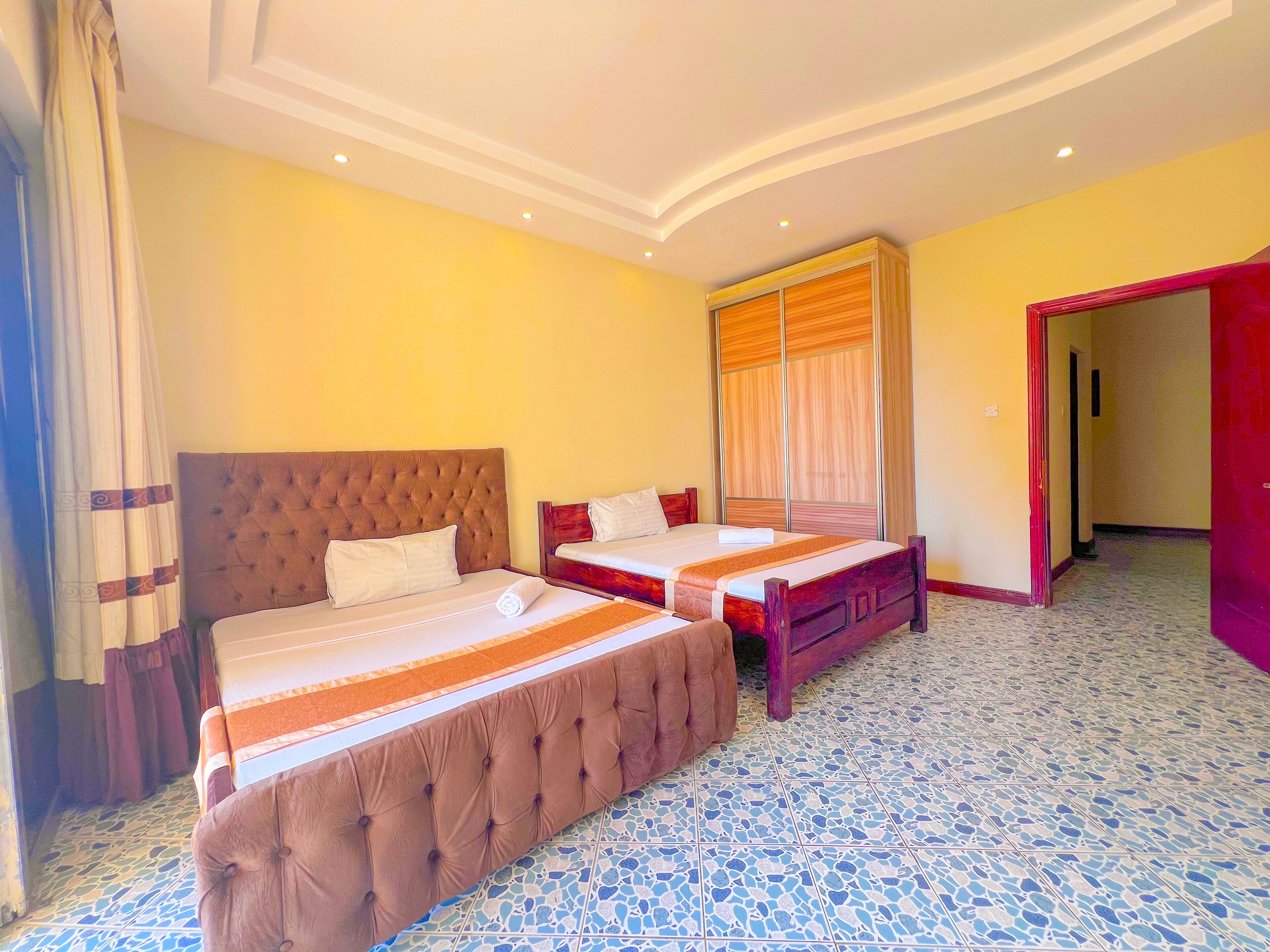 Quarto de hotel com duas camas, uma janela grande com cortinas, chão de azulejos e porta que dá para outro quarto.