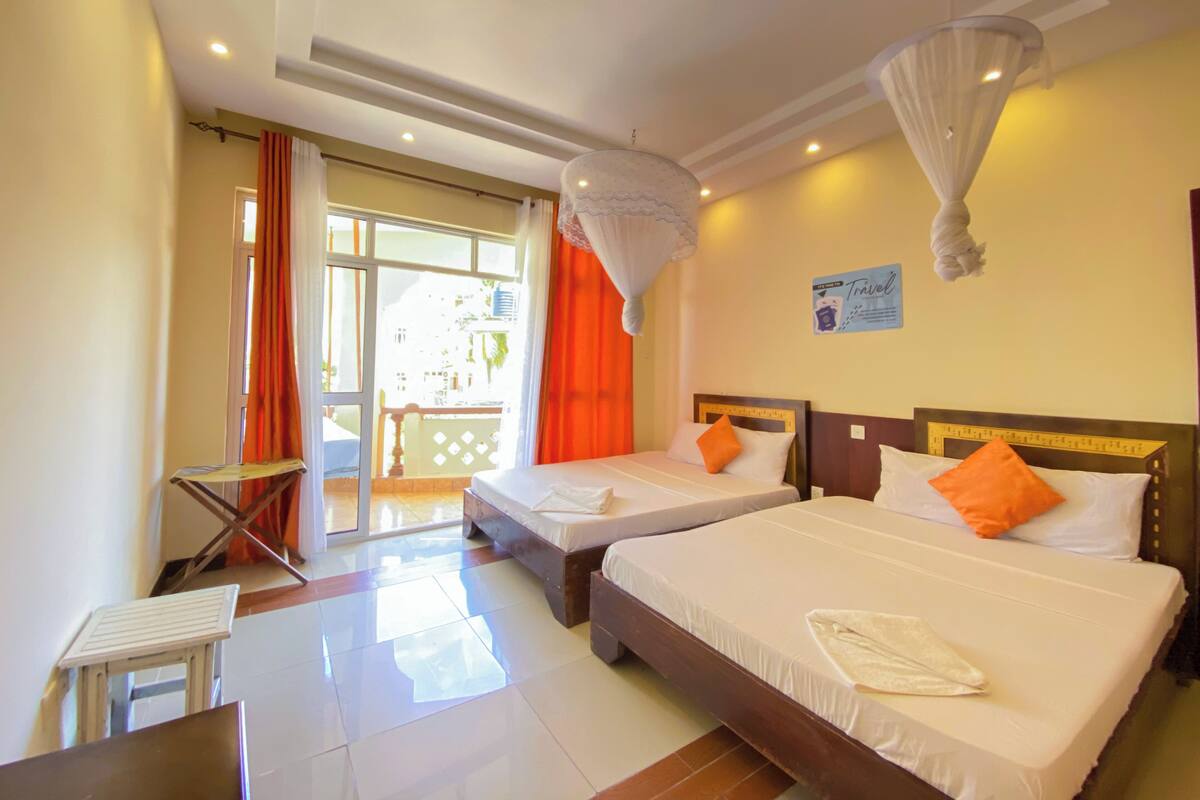 Une chambre d’hôtel avec deux lits, une petite table, une chaise et un balcon avec des rideaux orange.