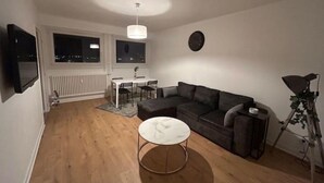 A living room with a sofa, a coffee table, a dining table with chairs, a TV mounted on the wall, and a window with a view of the city at night. - Une vue Imprenable sur Metz ! (Metz)