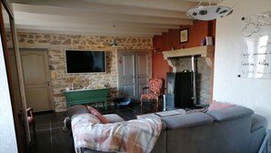 A living room with a grey sofa, a television, a fireplace, and a stone wall. - Maison Limousine en Pierre Apparentes (Compreignac)