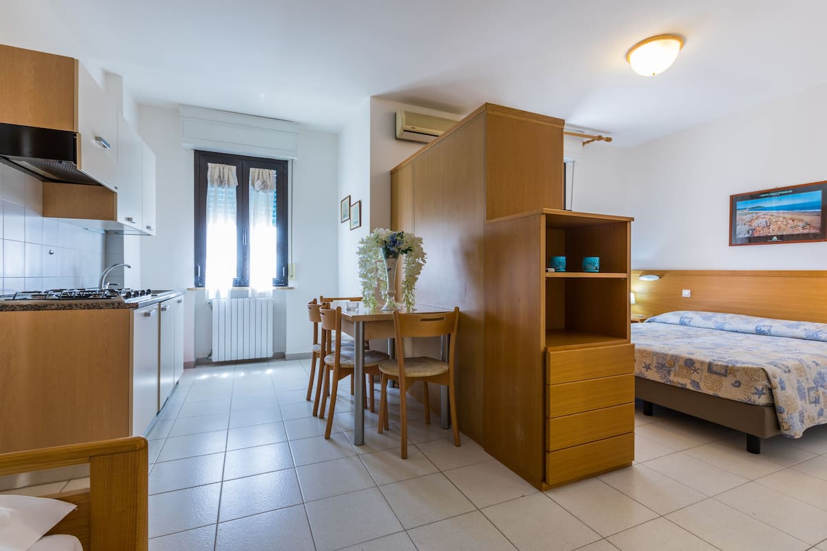 A compact room with a kitchenette, a dining area with a table and chairs, a bed with a patterned bedspread, and a wooden wardrobe.