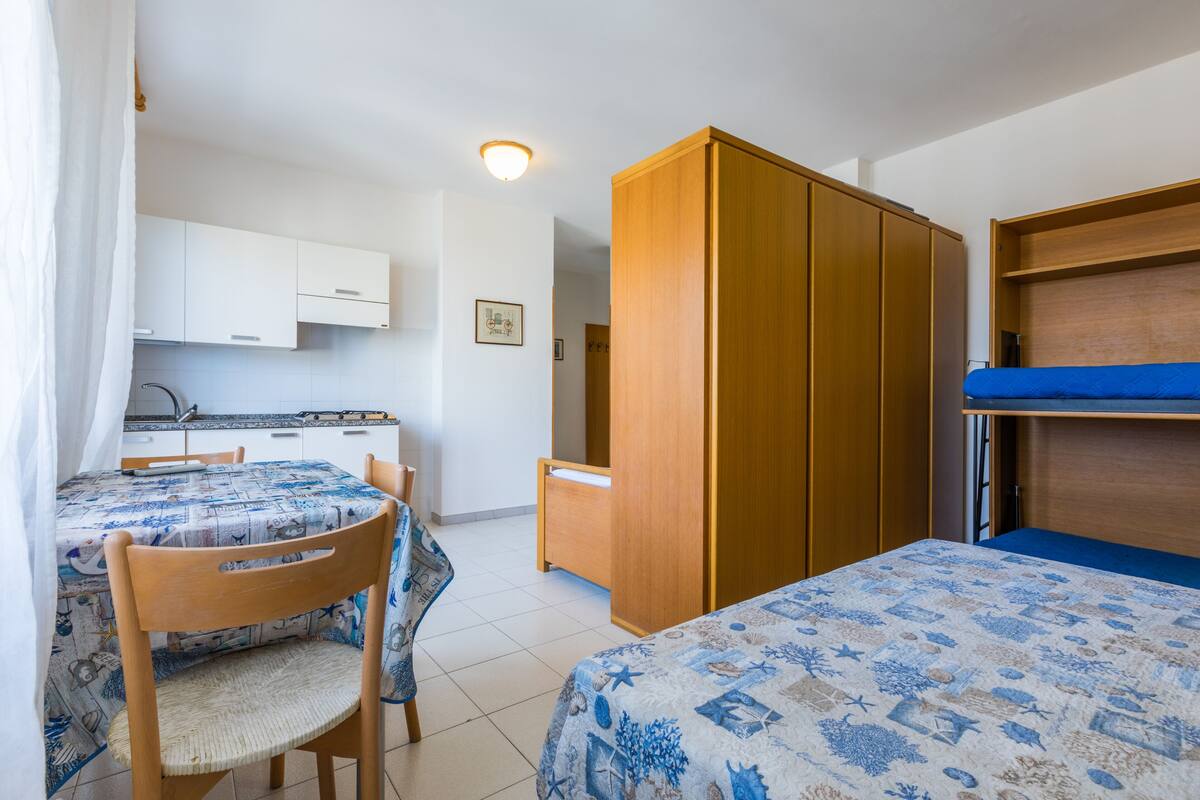 A room with a bunk bed, a dining table with chairs, a wooden wardrobe, and a kitchenette.