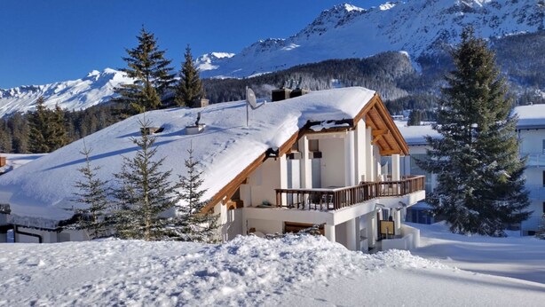 Una cabaña con balcón de madera, rodeada de montañas nevadas y pinos.