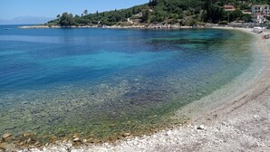 A coastal scene with clear waters, a pebble beach, and buildings on a hillside. - Solaris Methea Deluxe Suites  (Kassiopi)
