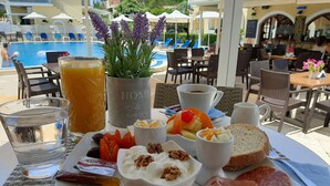 A breakfast tray with ham, cheese, fruit, and a drink, set on a table with a pool view in the background. - Solaris Methea Deluxe Suites  (Kassiopi)