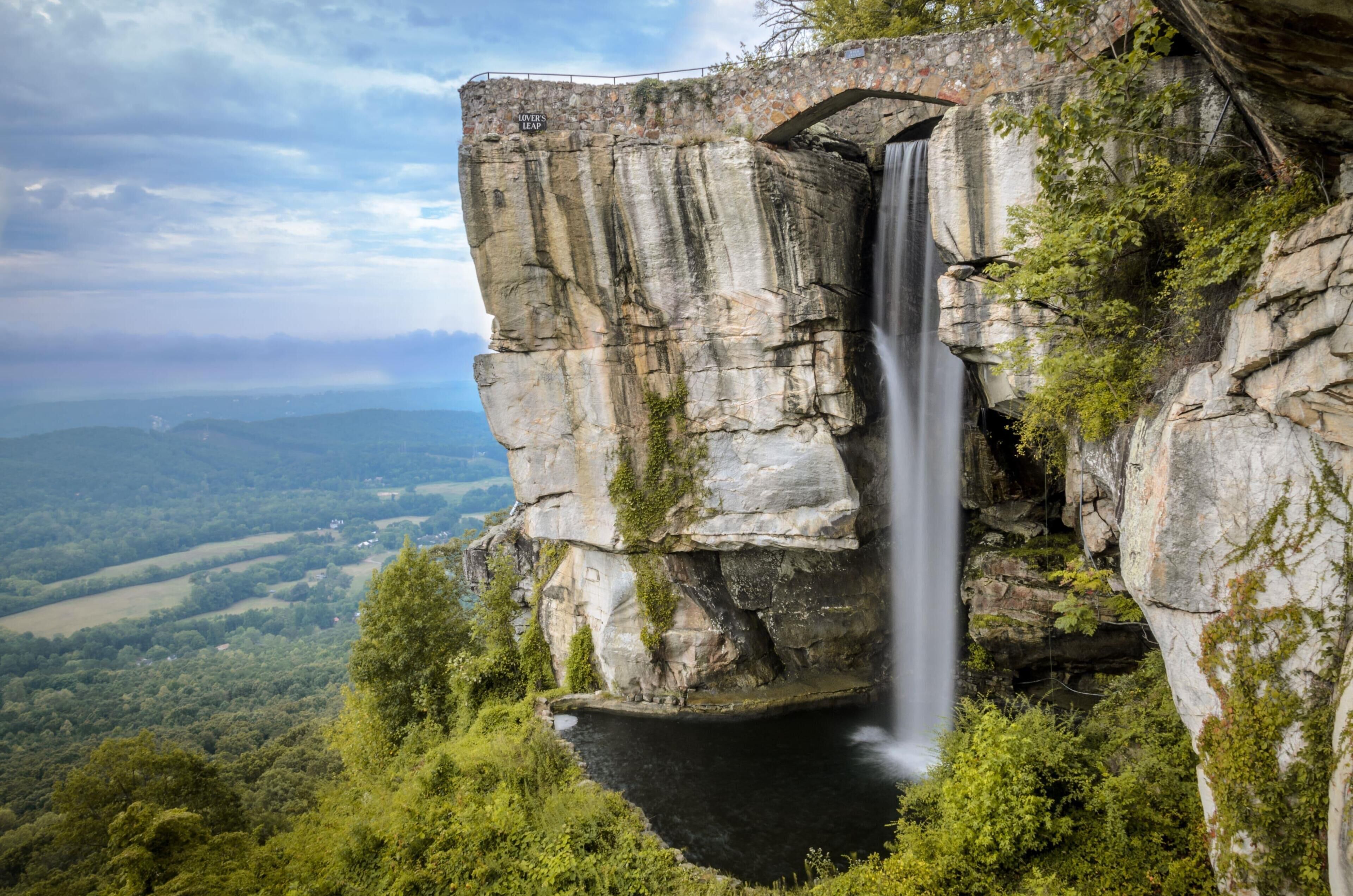 A waterfall cascading down a rocky cliff with a bridge and a view of the surrounding landscape.