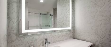 Un baño con pared de diseño marmolizado, un lavamanos blanco, un box de ducha de vidrio y un espejo con marco iluminado.