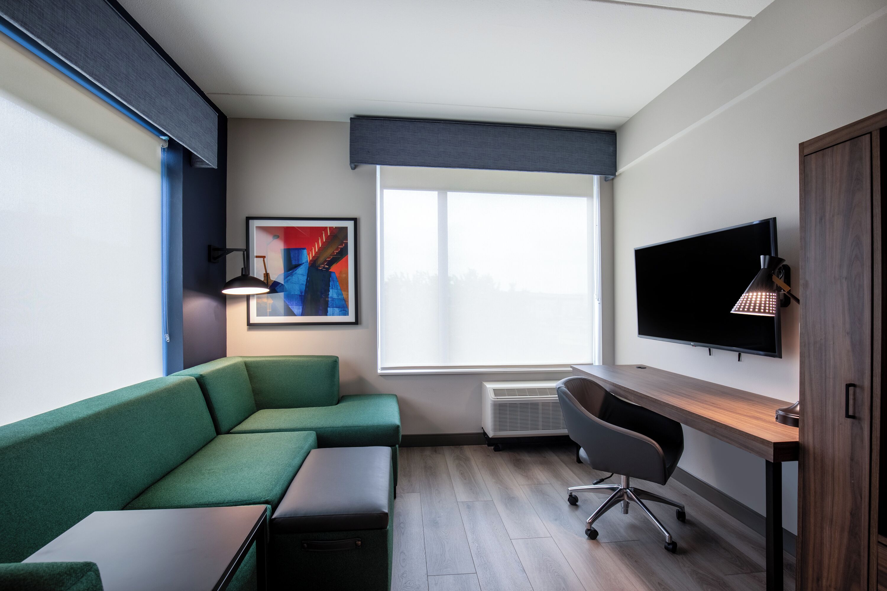 A modern hotel room with a green sofa, a desk, a chair, a flat-screen TV, and a large window.