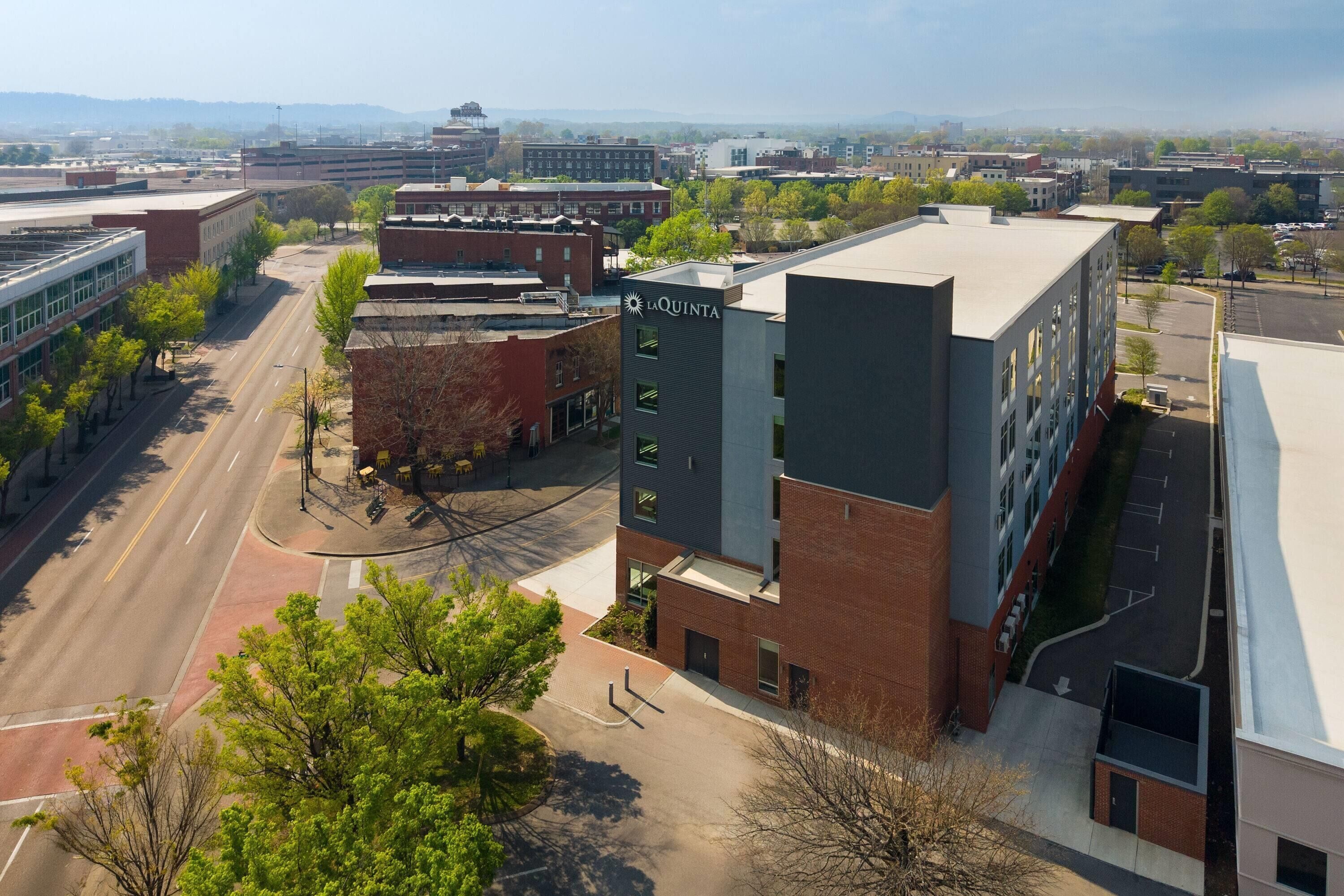 A multi-story building with a flat roof, a parking lot, and a road with trees and a clear sky in the background.
