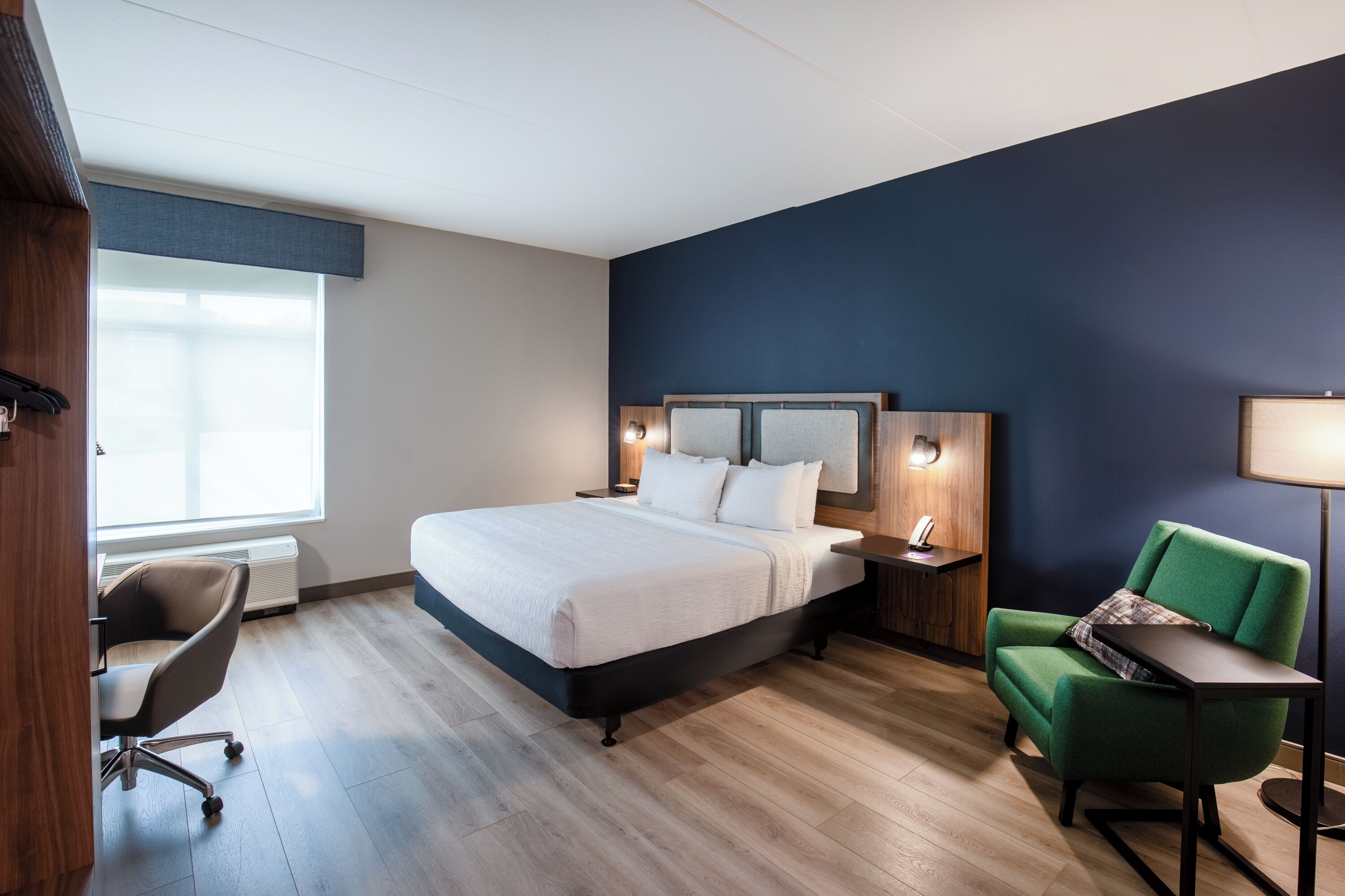 A modern hotel room with a large bed, a desk, a chair, and a lamp.