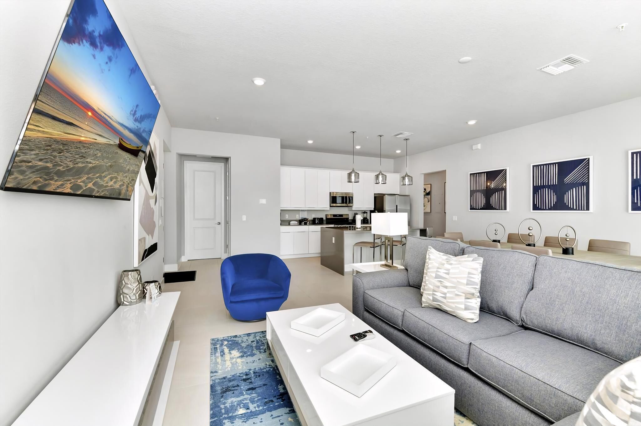 A modern living room with a grey sofa, a blue chair, and a white coffee table. There is a kitchen in the background with white cabinets and stainless steel appliances.
