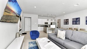 A modern living room with a grey sofa, a blue chair, and a white coffee table. There is a kitchen in the background with white cabinets and stainless steel appliances. - Tranquil Condo near Disney with Resort & Waterpark Access - B8-134 (Kissimmee)