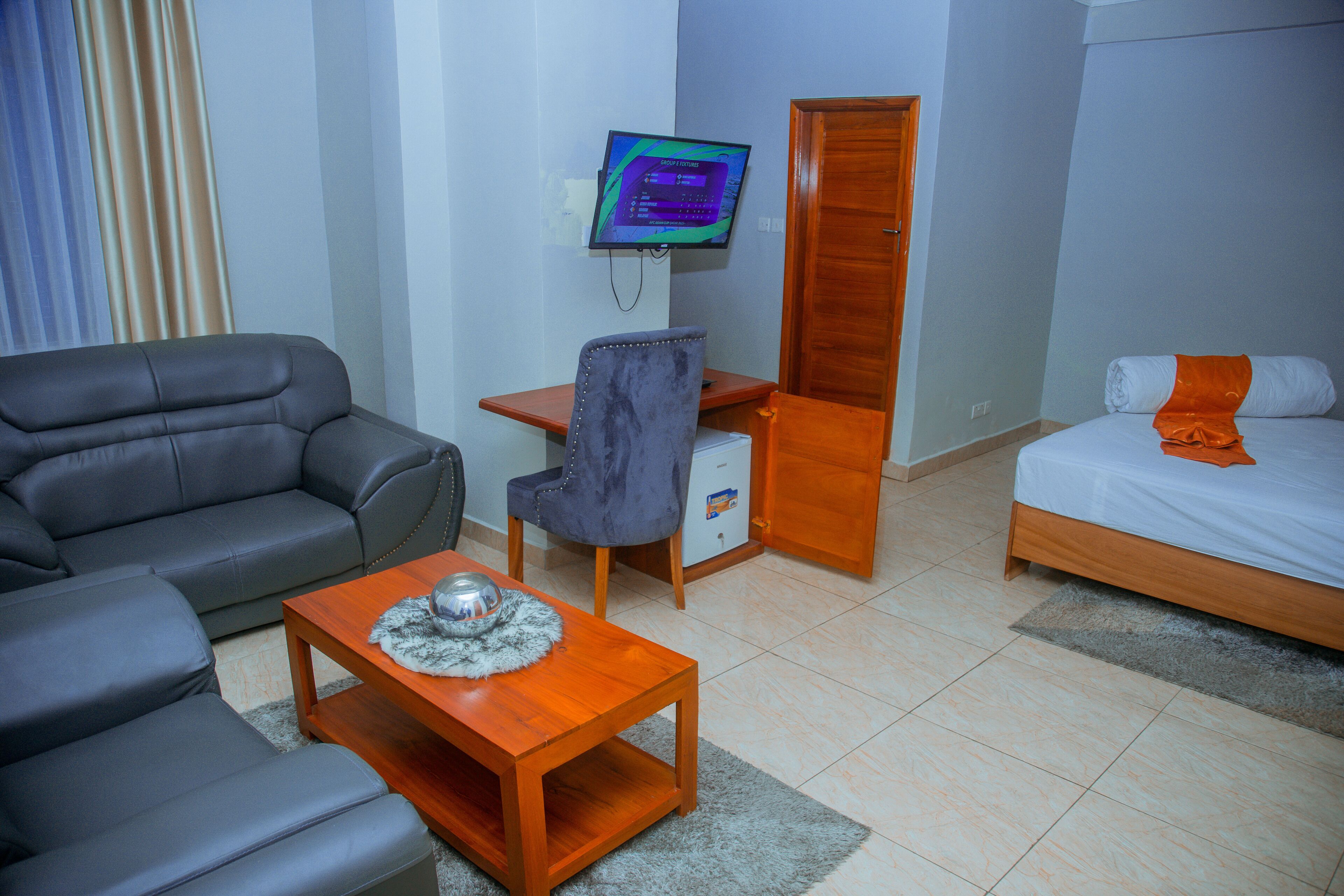 Une chambre d’hôtel avec un canapé, un bureau avec un ordinateur, un lit avec une couverture orange et une armoire en bois.