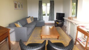 A living room with a grey sofa, a wooden coffee table, and two black armchairs. There is a television on a stand, a mirror above a fireplace, and a window with curtains. - Ontdek de Kracht van de Rust, Dichtbij de Kust! (Veurne)