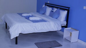 A neatly made bed with white linens and blue accents, a bedside table, and a small rug on a tiled floor. - ELAND ACCOMMODATIONS (Ongata Rongai)