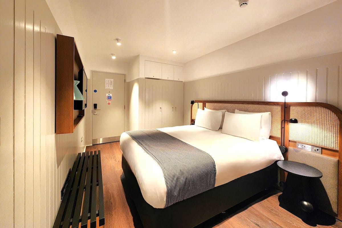 A modern hotel room with a large bed, a wooden bench, a small table, and a lamp.