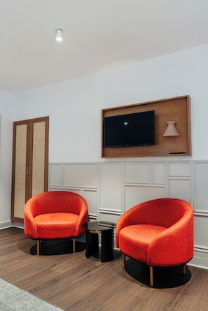 Quarto com duas poltronas vermelhas, uma mesinha preta, uma televisão presa a um painel de madeira e uma porta com painel trançado.