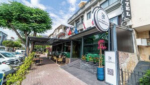 Un hotel con un letrero que dice "Deniz Boutique Hotel" y un código QR. Hay mesas y sillas al aire libre, y una planta en maceta con flores rosas.