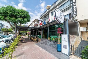 Un hotel con un letrero que dice "Deniz Boutique Hotel" y un código QR. Hay mesas y sillas al aire libre, y una planta en maceta con flores rosas.