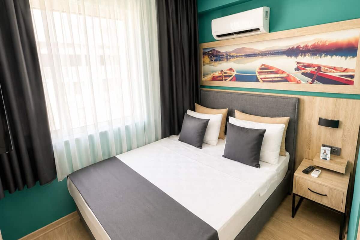 A hotel room with a bed, a nightstand, an air conditioning unit, a painting, and a window with curtains.