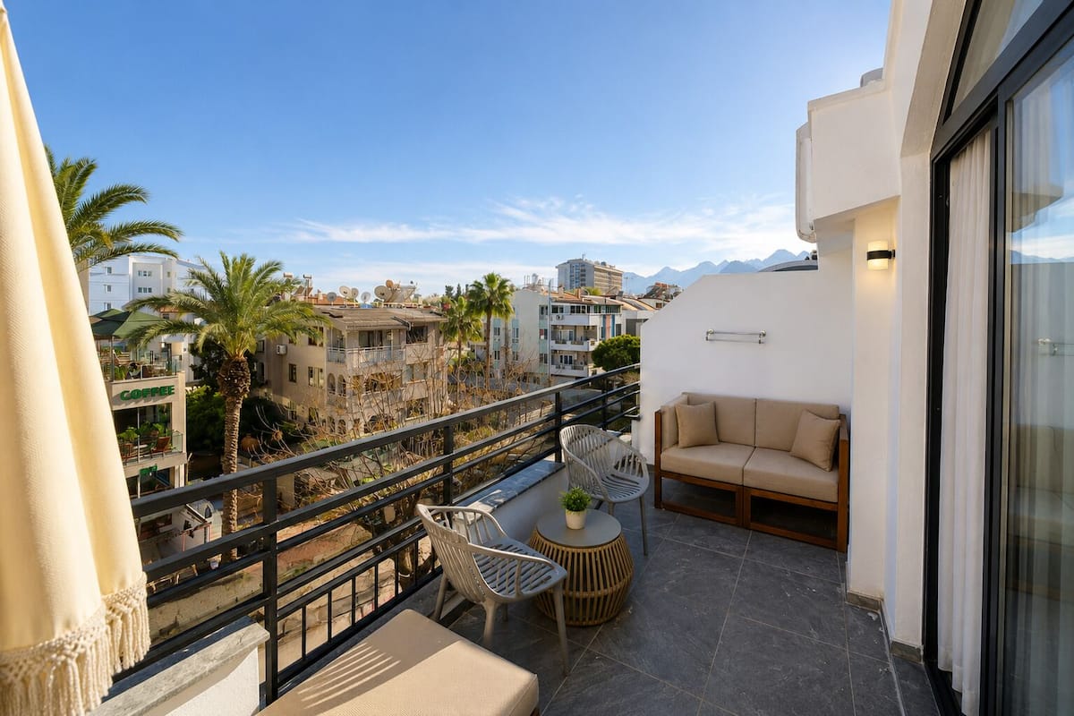 A balcony with a sofa, chairs, and a table, overlooking a cityscape with buildings and palm trees.