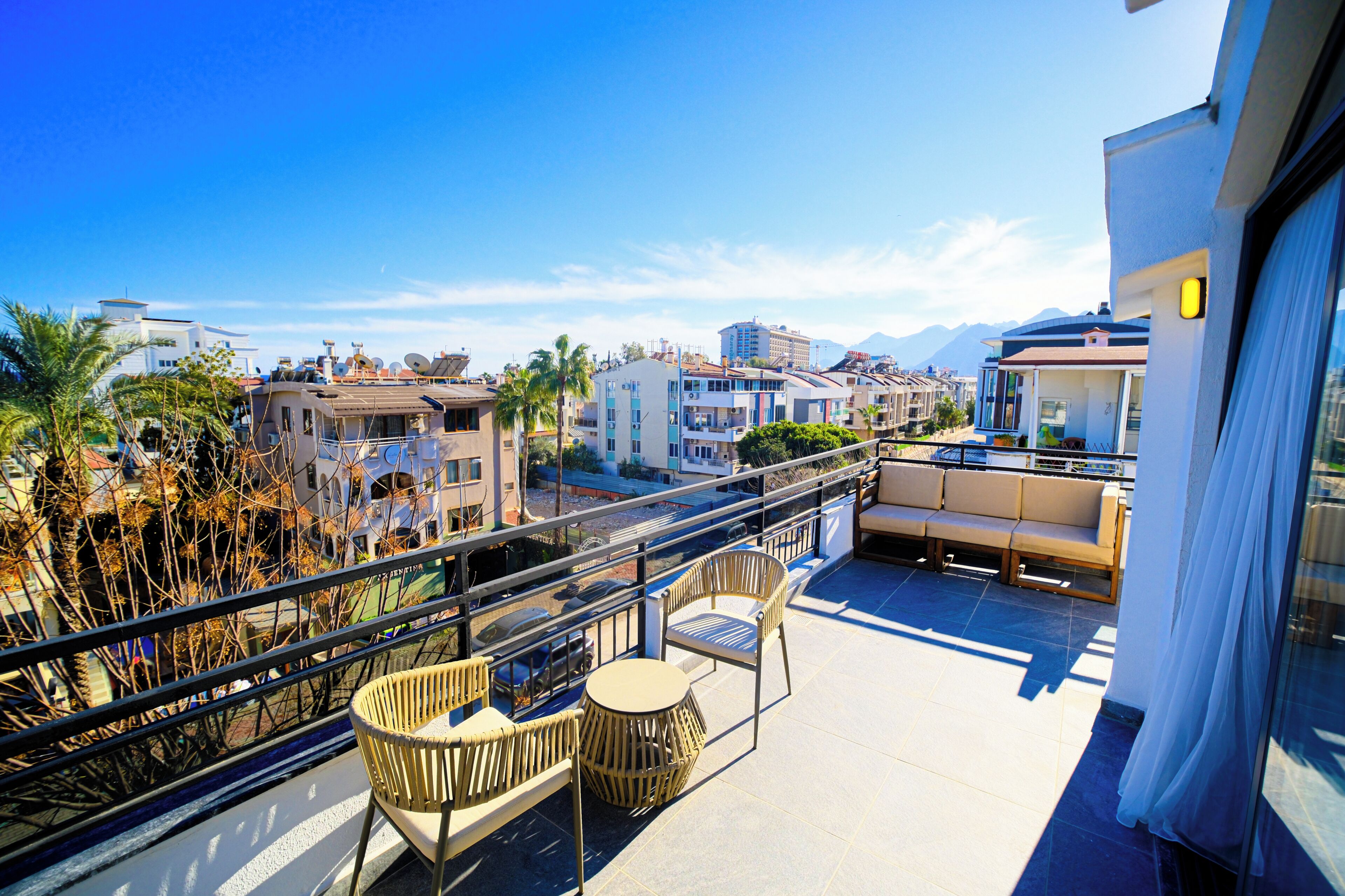 A balcony with wicker furniture, a glass railing, and a view of surrounding buildings and mountains.