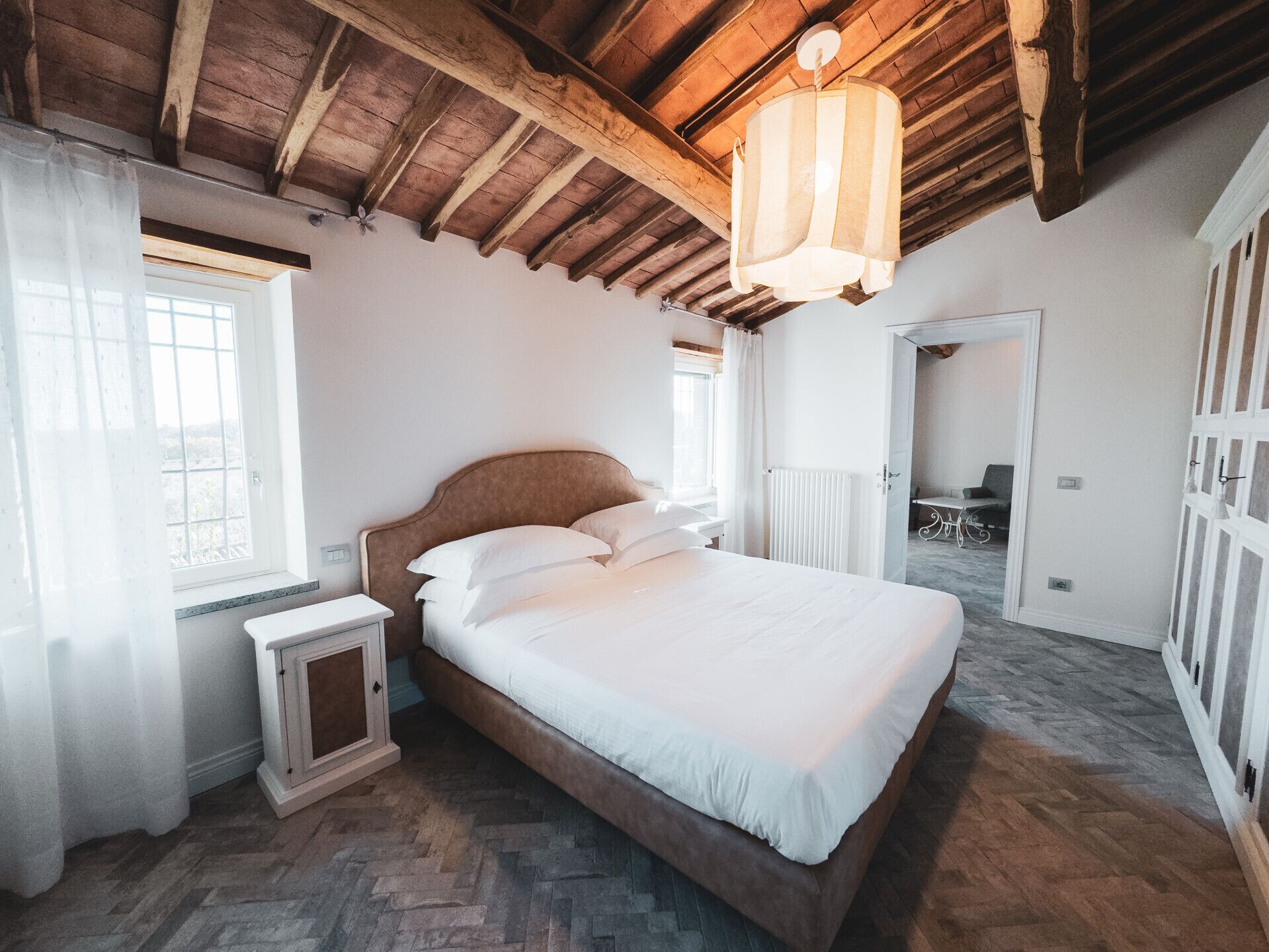Un dormitorio con techo de madera, una cama con sábanas blancas, una mesita de noche y una ventana con cortinas.