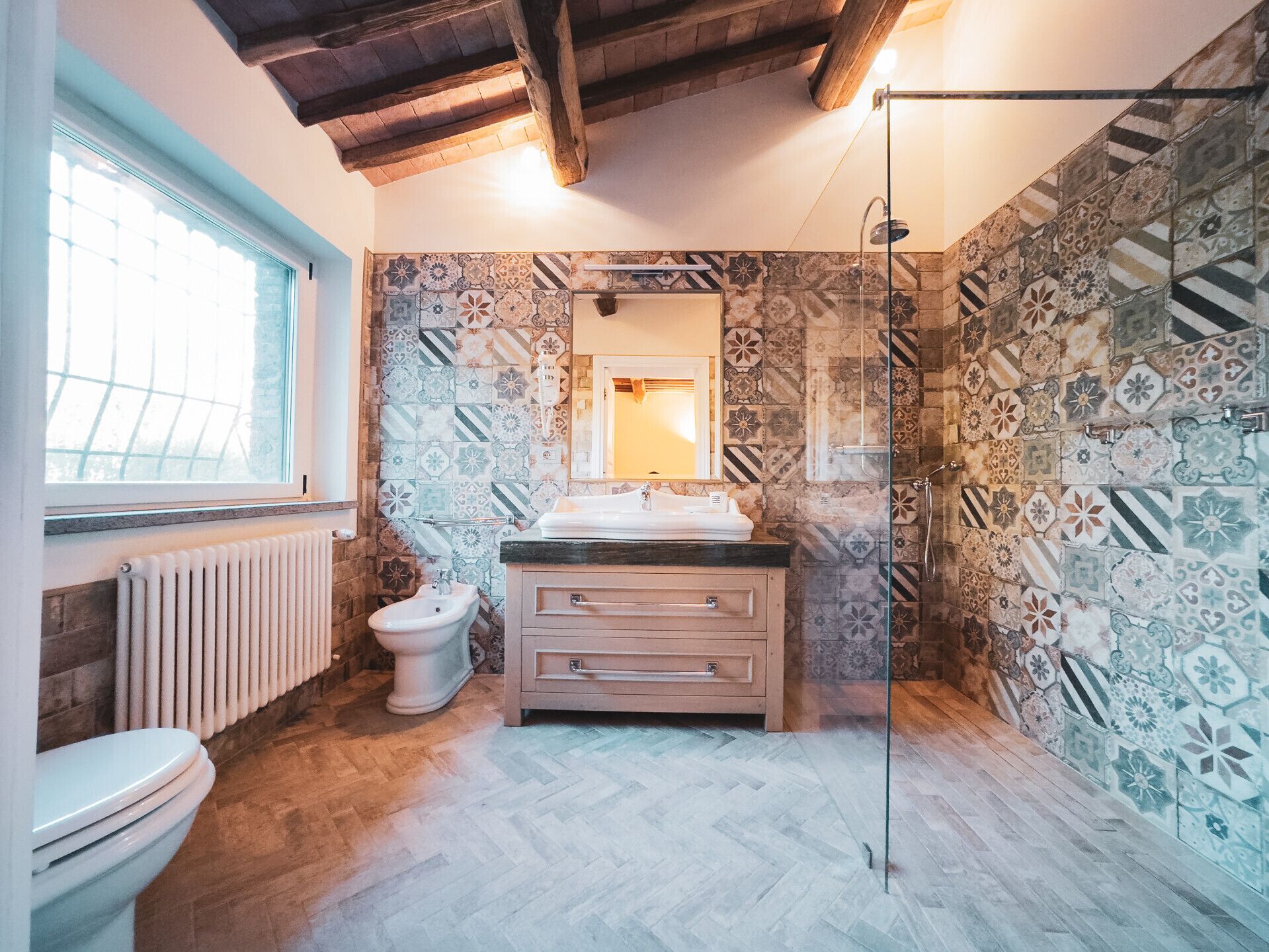 Een badkamer met een visgraat vloerpatroon, een wit toilet, een wastafel met spiegel en een douche met glazen deur.