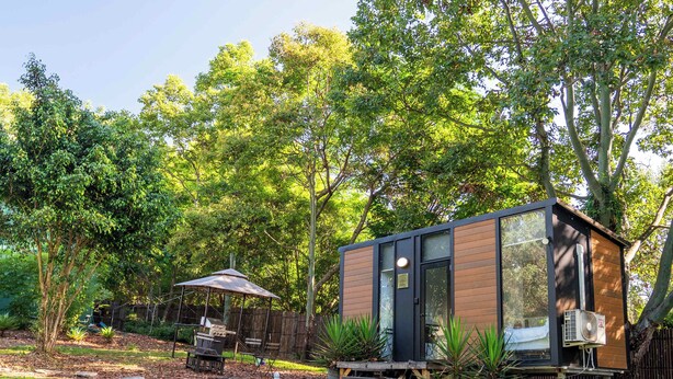 Une petite maison moderne, avec une façade en briques et de grandes fenêtres, entourée d’arbres et d’une pelouse.