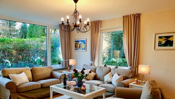 A well-lit living room with two sofas, a coffee table, and a chandelier. Large windows offer views of greenery outside. - fewo1846 - Kranich Museumsberg - apartment with 2 bedrooms on the ground floor (Flensburg)