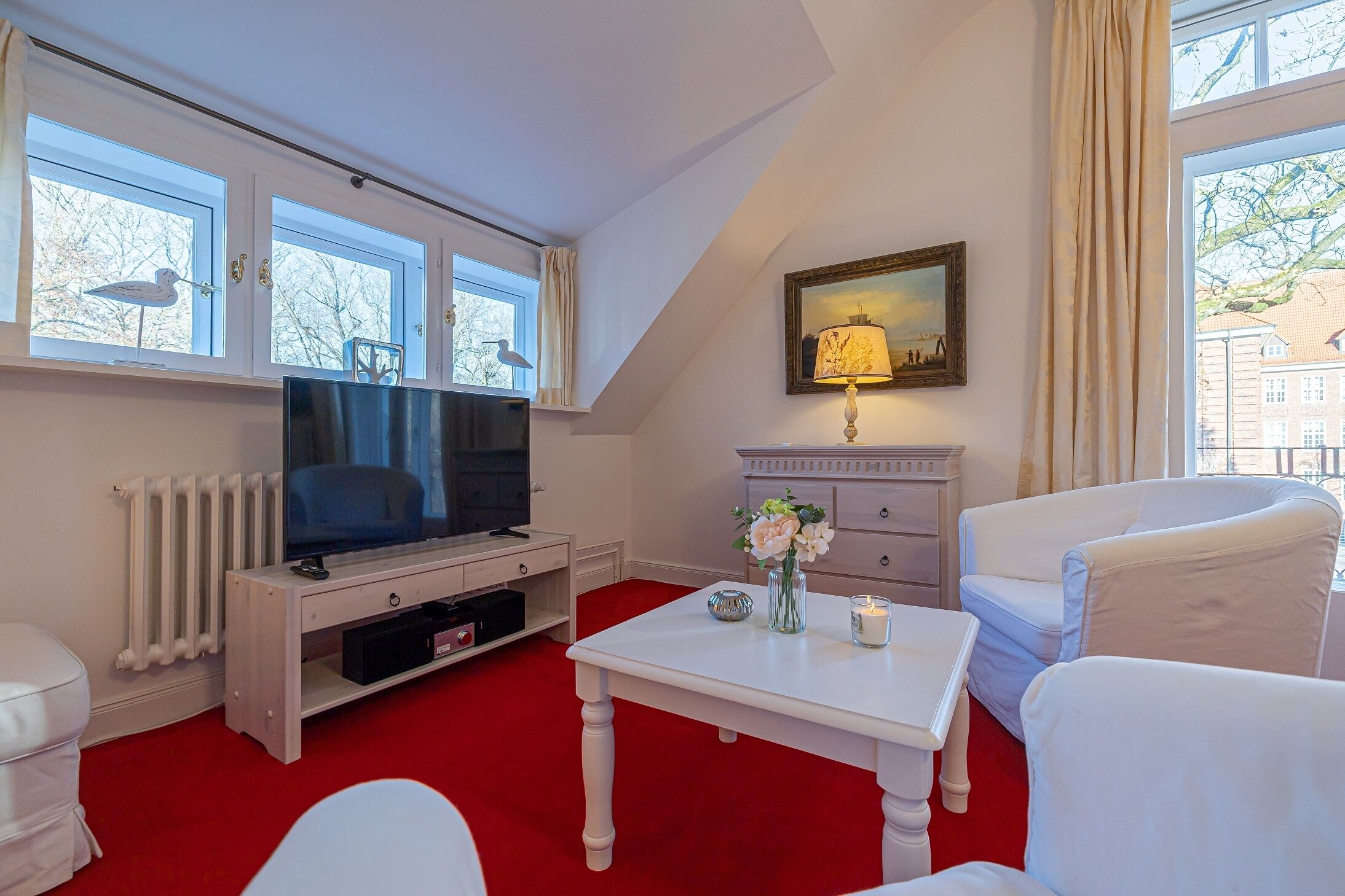 A room with a television on a white stand, a white table with a vase of flowers, a lamp, and a chair.