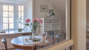 A dining area with a round table, chairs, a vase with flowers, and a window with blinds. - fewo1846 - Villa Royal (Flensburg)