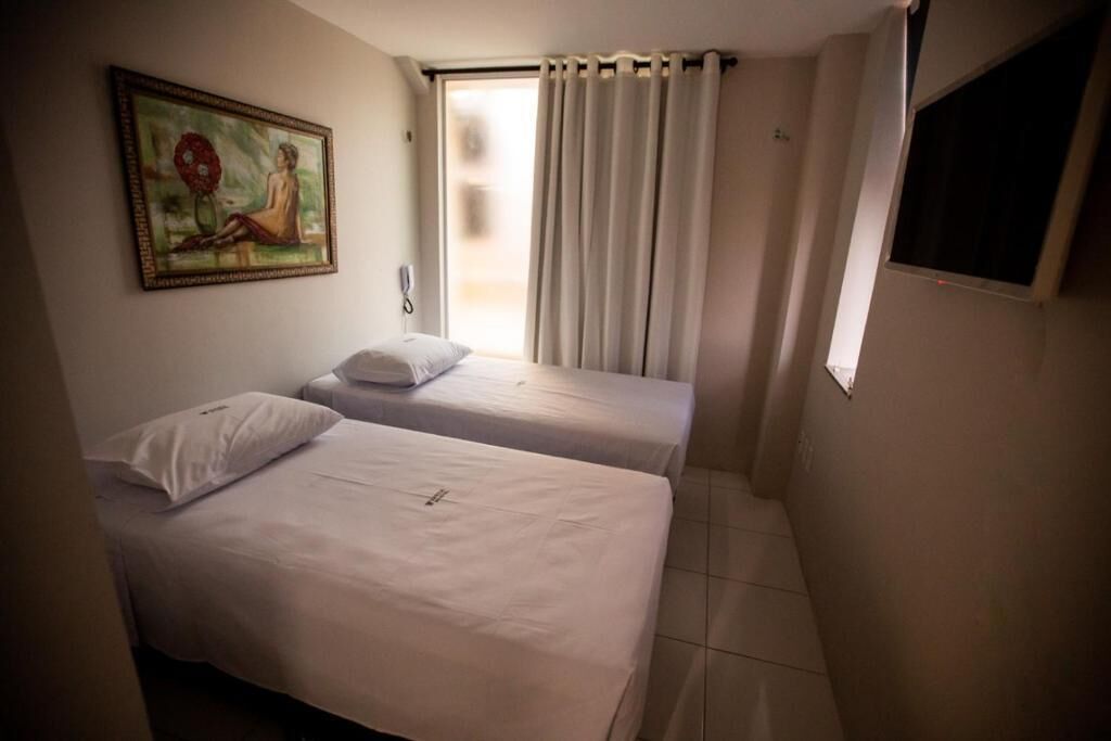 A small, clean room with two single beds, a painting on the wall, and a window with curtains.