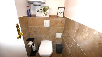 Ein kleines Badezimmer mit Toilette, Mülleimer, Toilettenpapierhalter und einem gerahmten Bild an der Wand.
