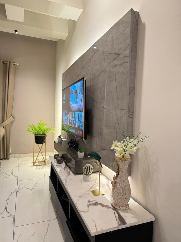 Uma sala de estar moderna com uma grande televisão de tela plana fixada em uma parede de mármore, piso de ladrilho de mármore, um aparador com objetos decorativos e um vaso de plantas.