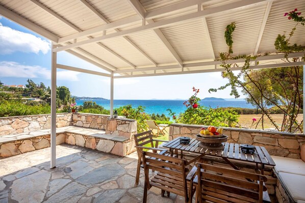 A covered outdoor seating area with stone walls, wooden furniture, and a view of the sea.