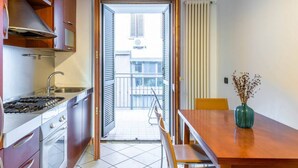 Een kleine keuken met een kookplaat, spoelbak en kastjes, die uitkomt op een balkon met uitzicht op gebouwen.