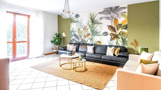 Een woonkamer met een zwarte leren bank, een ronde salontafel en een groot tropisch muurdecor.