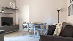 Una sala moderna con un sofá gris, una mesa de comedor con sillas, un televisor de pantalla plana sobre un mueble bajo y una lámpara de pie.