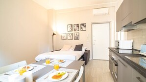 Una habitación de hotel moderna con cama, zona de comedor con desayuno, kitchenette y obras de arte en la pared.
