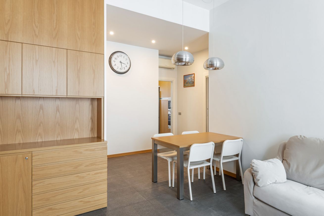 Una habitación moderna con un armario de madera, una mesa de comedor con sillas, un sofá blanco, un reloj y lámparas colgantes.