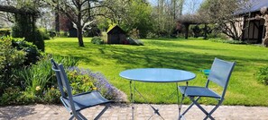 Un jardin avec une table et deux chaises, entouré d’une végétation luxuriante et d’un petit bâtiment en briques.
