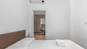 A modern hotel room with a neatly made bed, a small bedside table, and a view into the adjacent room.