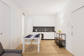 Cozinha compacta com armários brancos, painel de fundo preto e área de jantar com mesa e cadeiras brancas.