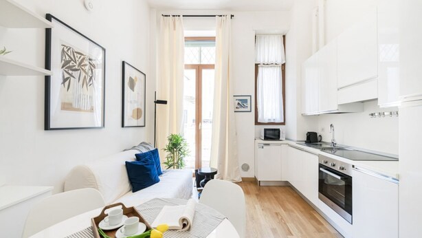 A compact kitchen with white cabinets, a microwave, and a stove. There is a dining area with a table set for two, and a small window with curtains. - Easylife - Magic flat in Duomo (Milano)