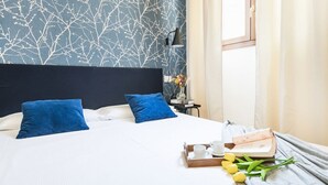 A neatly made bed with blue pillows, a bedside table with a lamp, a tray with a book and a cup, and a window with curtains. - Easylife - Magic flat in Duomo (Milano)